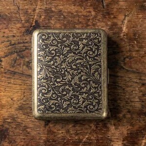 May include: A rectangular metal case with a dark brown finish and an embossed gold-toned floral scrollwork pattern covering the entire front surface. The edges of the case feature a smooth, rounded gold-toned metal frame with a visible side hinge.