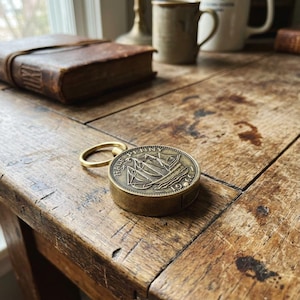 May include: A vintage brass Half Penny coin with a ship design, attached to a gold-colored keyring. The coin is set in a small, round case. The coin is on a rustic wooden table.