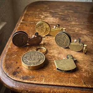 May include: A collection of vintage brass lighters and a keychain, featuring ship designs on the coins. The lighters have a rectangular shape and a hinged lid. The keychain is round with a ship engraving. The items are on a wooden surface.