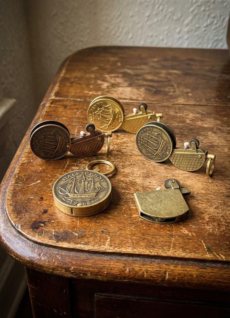 May include: A collection of vintage brass lighters and a keychain, featuring ship designs on the coins. The lighters have a rectangular shape and a hinged lid. The keychain is round with a ship engraving. The items are on a wooden surface.