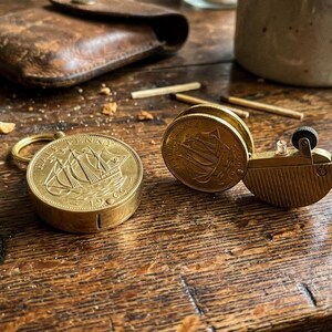 May include: A vintage brass match safe and lighter set. The match safe features a ship engraving on the coin-shaped lid. The lighter has a similar coin design. The items are on a wooden surface.