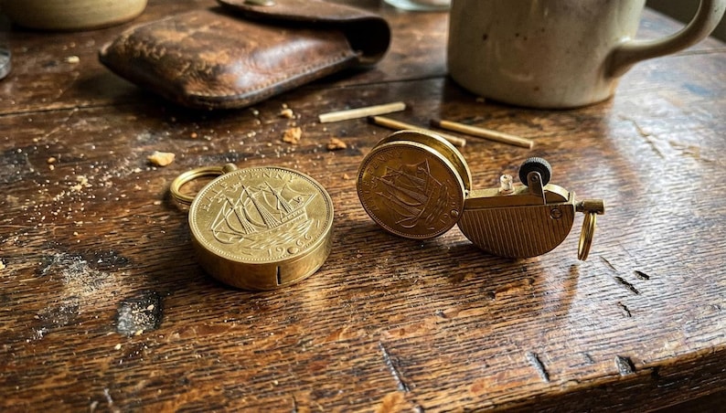 May include: A vintage brass match safe and lighter set. The match safe features a ship engraving on the coin-shaped lid. The lighter has a similar coin design. The items are on a wooden surface.