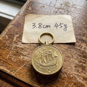 May include: A gold-colored Half Penny coin from 1966, featuring a sailing ship, attached to a keyring. The coin measures 1.5 inches in diameter and weighs 1.6 ounces. A handwritten note indicates the coin's dimensions.