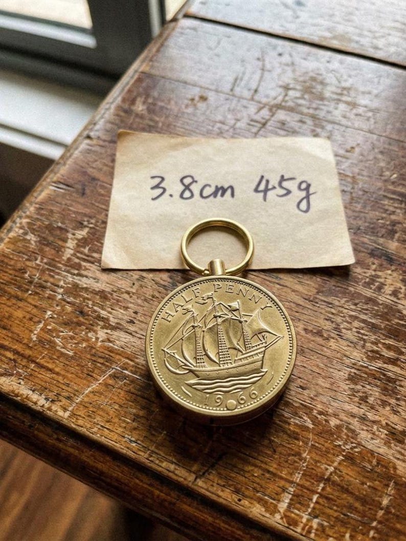May include: A gold-colored Half Penny coin from 1966, featuring a sailing ship, attached to a keyring. The coin measures 1.5 inches in diameter and weighs 1.6 ounces. A handwritten note indicates the coin's dimensions.