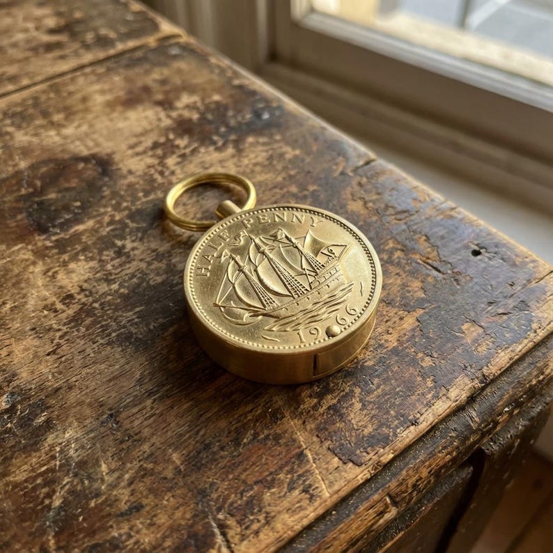 May include: A gold-colored, round keychain featuring a 1966 Half Penny coin with a sailing ship design. The coin is attached to a small, circular case with a gold ring for attaching keys. The keychain rests on a weathered, wooden surface.