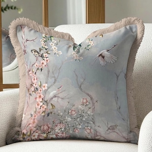 May include: Square decorative pillow with a floral and bird design. The pillow features a light blue background with pink flowers, birds, and tree branches. The edges are trimmed with a beige fringe.