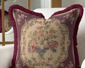 May include: A decorative throw pillow with a floral design. The pillow features a central bouquet of pink, purple, and blue flowers on a beige background, framed by a burgundy border and fringe.