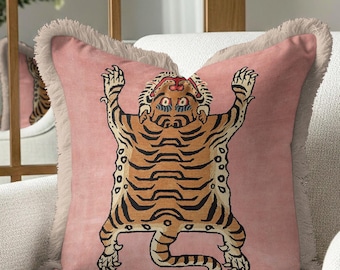 May include: A decorative pink throw pillow with a fringed edge. The pillow features a stylized tiger design in shades of brown and black. The tiger is depicted with its arms raised. The pillow is placed on a white armchair.