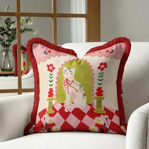 May include: Decorative square pillow featuring a whimsical cat design. The pillow has a red and pink checkered pattern, a white cat with floral accents, and a red fringe border. The cat is inside a green archway with floral accents.