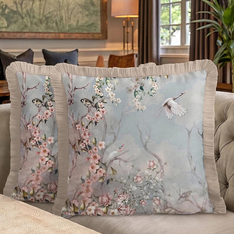 May include: Two decorative throw pillows with a floral and bird design. The pillows feature a light blue background with pink flowers, birds, and a flying crane. The edges are trimmed with a beige ruffle.
