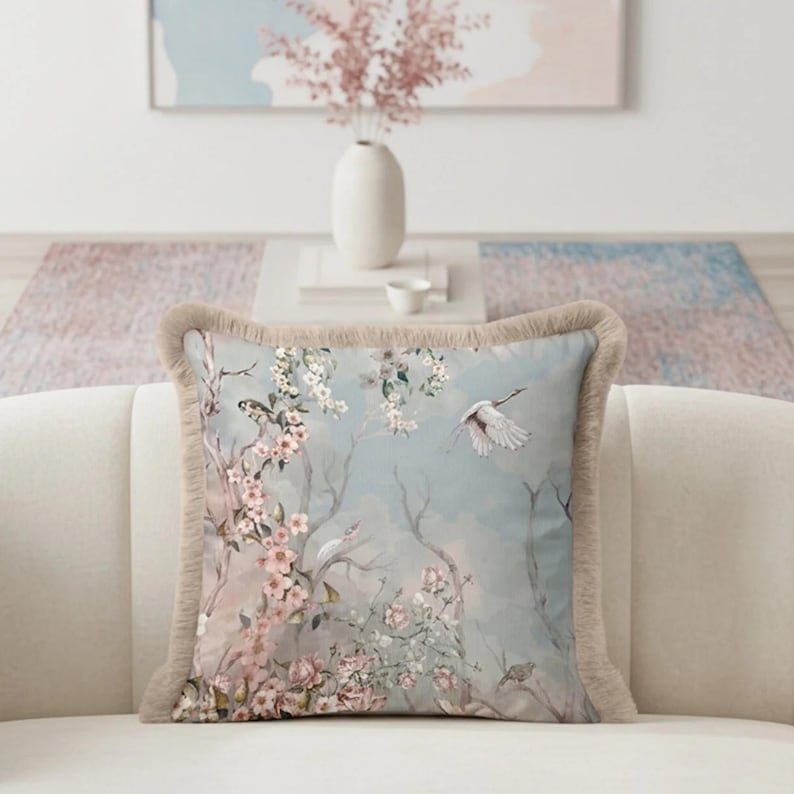 May include: Square decorative pillow with a floral and bird design. The pillow features a light blue background with pink cherry blossoms, birds, and a flying crane. The pillow has a beige fringe border.