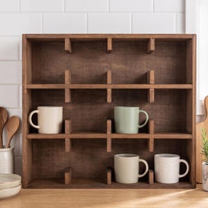 May include: A dark brown wooden display shelf featuring a grid of twelve open compartments. Four ceramic mugs in cream, sage green, light beige, and white with speckles are placed within the individual cubbies of the wooden organiser unit.