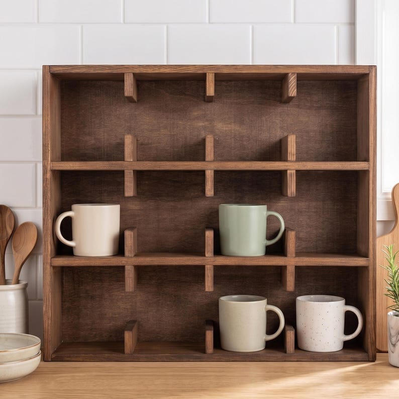 May include: A dark brown wooden wall-mounted display shelf with twelve square compartments. Four ceramic mugs in cream, sage green, light beige, and white with speckles are placed inside the individual cubbies of the wooden organizer unit.