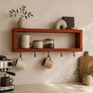 May include: A rectangular wooden wall shelf with a rich brown finish, featuring five black metal hooks underneath. The shelf holds two stacked beige mugs, a beige canister, a glass jar with coffee beans, a small vase with greenery, and a book.
