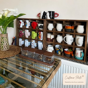 May include: A dark brown wooden grid shelf holding various ceramic mugs with different colors, patterns, and shapes. A set of four square glass jars with wooden lids sits in front. A vase with white calla lilies is on the left side.