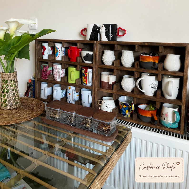 May include: A dark brown wooden grid shelf holding various ceramic mugs with different colors, patterns, and shapes. A set of four square glass jars with wooden lids sits in front. A vase with white calla lilies is on the left side.