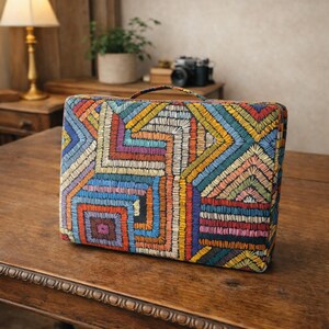 Puede incluir: Un bolso rectangular bordado y colorido con asa. El bolso presenta un patr&oacute;n geom&eacute;trico en tonos azules, amarillos, rojos y morados. El bolso est&aacute; sobre una mesa de madera.