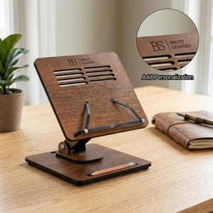 May include: A wooden book stand with the text "BS Brody Sanders" and the words "Add Personalization". The stand is brown with black metal supports and a pencil holder. A notebook and pen are on the table.