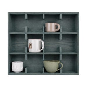 May include: A dark blue-gray wooden wall shelf with twelve square compartments. Four mugs are placed inside: a solid light pink mug, a white mug with a small brown graphic, a white mug with a sculpted face design, and a solid light green mug.