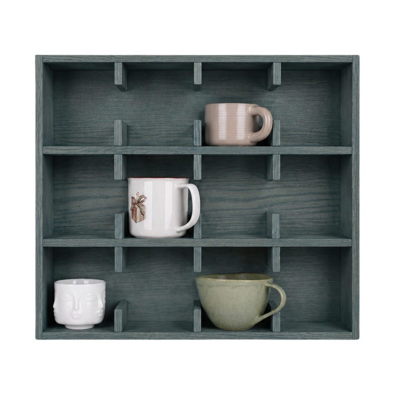 May include: A dark blue-gray wooden wall shelf with twelve square compartments. Four mugs are placed inside: a solid light pink mug, a white mug with a small brown graphic, a white mug with a sculpted face design, and a solid light green mug.