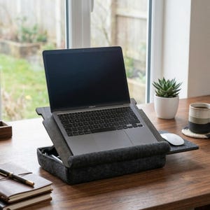 Op de afbeelding: Een laptop staat op een grijze vilten laptopstandaard met een ingebouwd opbergvak. Een witte muis en een kleine vetplant in een witte pot staan ook op het houten bureau.
