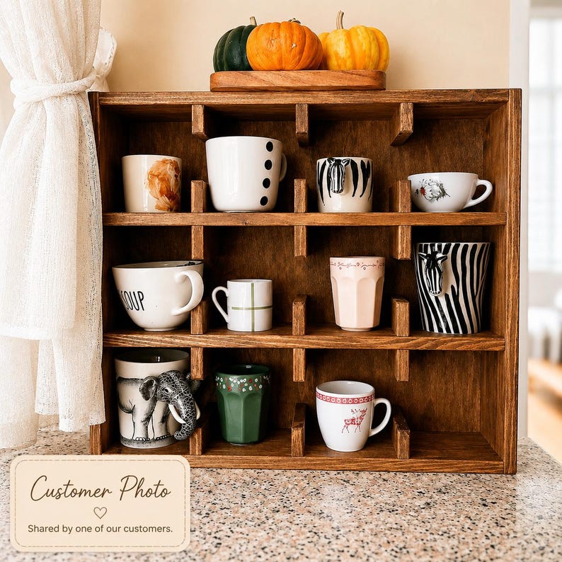 May include: A dark brown wooden display shelf with twelve square compartments holding various ceramic mugs. The mugs feature diverse designs including animal motifs, patterns, and text. Three small pumpkins sit on top of the wooden unit.