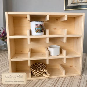 May include: A light-colored wooden display shelf with three horizontal tiers and vertical dividers. A white mug with a colorful Prague illustration, a plain white ceramic bowl, and a brown and white checkered ceramic mug are inside.