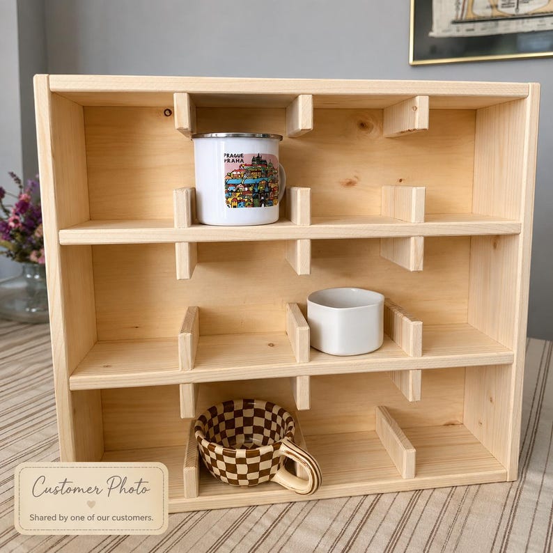 May include: A light-colored wooden display shelf with three horizontal tiers and vertical dividers. A white mug with a colorful Prague illustration, a plain white ceramic bowl, and a brown and white checkered ceramic mug are inside.