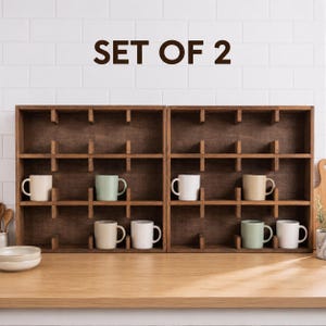 May include: A set of two dark brown wooden wall-mounted display shelves with multiple square compartments. The text SET OF 2 is displayed above the shelves. Several solid-colored ceramic mugs in white, cream, and light green are placed inside.