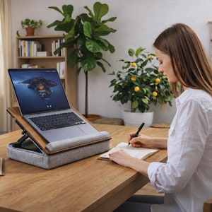 Op de afbeelding: Een laptop op een houten standaard met een grijs kussen, geplaatst op een houten bureau. Het laptopscherm toont een landschapsafbeelding. Een persoon schrijft met een pen in een notitieboek. Planten en boeken zijn zichtbaar op de achtergrond.