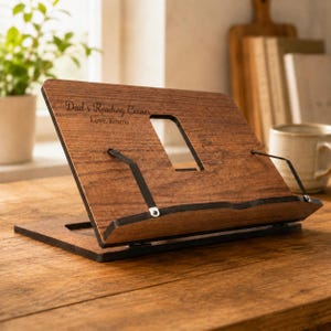 May include: A wooden book stand with the text "Dad's Reading Corner, Love, Emma". The stand is brown with black metal supports and a rectangular cutout. It is positioned on a wooden surface.