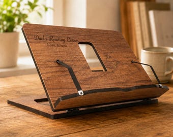 May include: A wooden book stand with the text "Dad's Reading Corner, Love, Emma". The stand is brown with black metal supports and a rectangular cutout. It is positioned on a wooden surface.