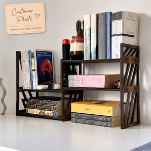 May include: A dark brown wooden desktop bookshelf with a tiered, open-frame design featuring diagonal side slats. The shelf holds various hardcover and paperback books, a small potted cactus, and a black cylindrical container on its surfaces.