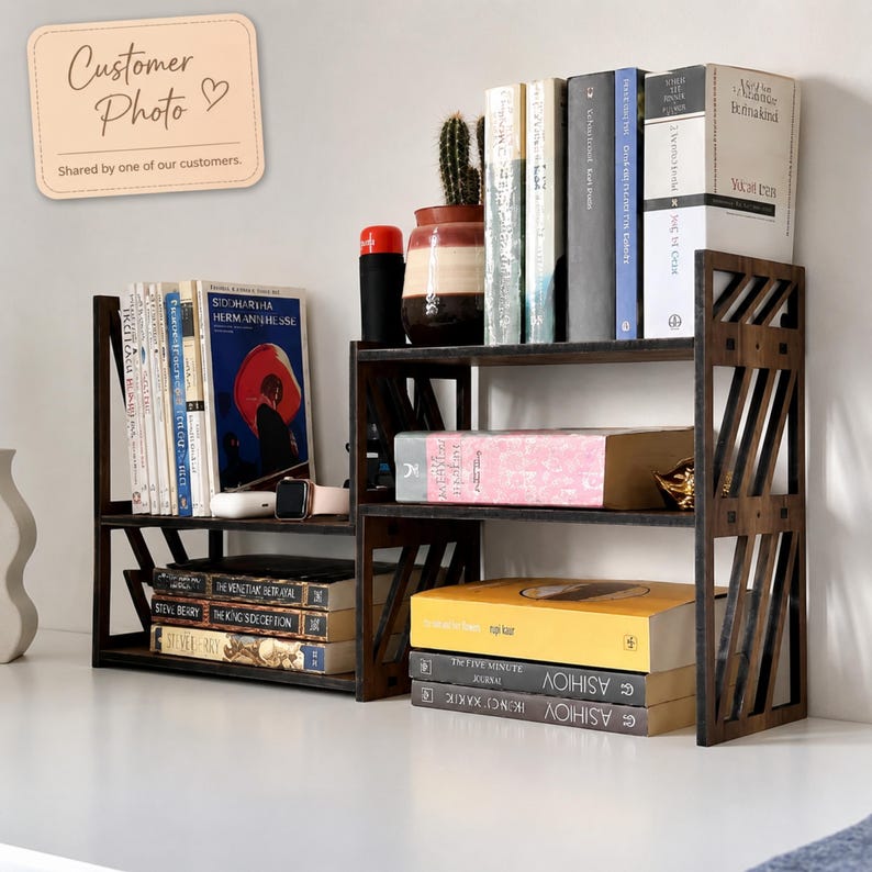 May include: A dark brown wooden desktop bookshelf with a tiered, open-frame design featuring diagonal side slats. The shelf holds various hardcover and paperback books, a small potted cactus, and a black cylindrical container on its surfaces.
