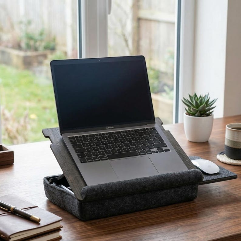 May include: A dark gray felt lap desk with a wooden laptop stand attachment. The stand holds a silver laptop with a black keyboard and trackpad. A white computer mouse sits on the right side of the felt base, which features a raised front edge.