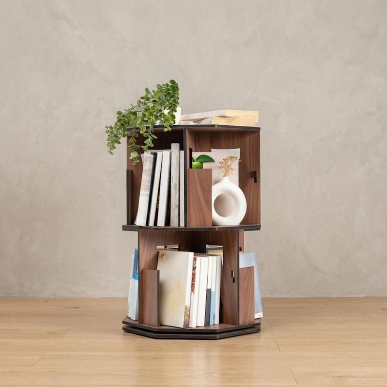 May include: A three-tiered, rotating bookshelf made of dark wood. The shelves are filled with books, a small white vase, and a potted plant. The bookshelf is designed to maximize space and storage.
