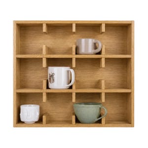 May include: A square light brown wooden wall shelf with twelve individual compartments. Four ceramic mugs are placed inside: a beige mug, a white mug with a small graphic, a white mug with a sculpted face design, and a sage green rounded mug.