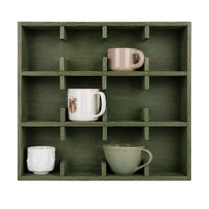 May include: A square, dark green wooden wall shelf with twelve individual compartments. Four mugs are placed inside: a light pink mug, a white mug with a small brown graphic, a white mug with a sculpted face design, and a sage green ceramic bowl.