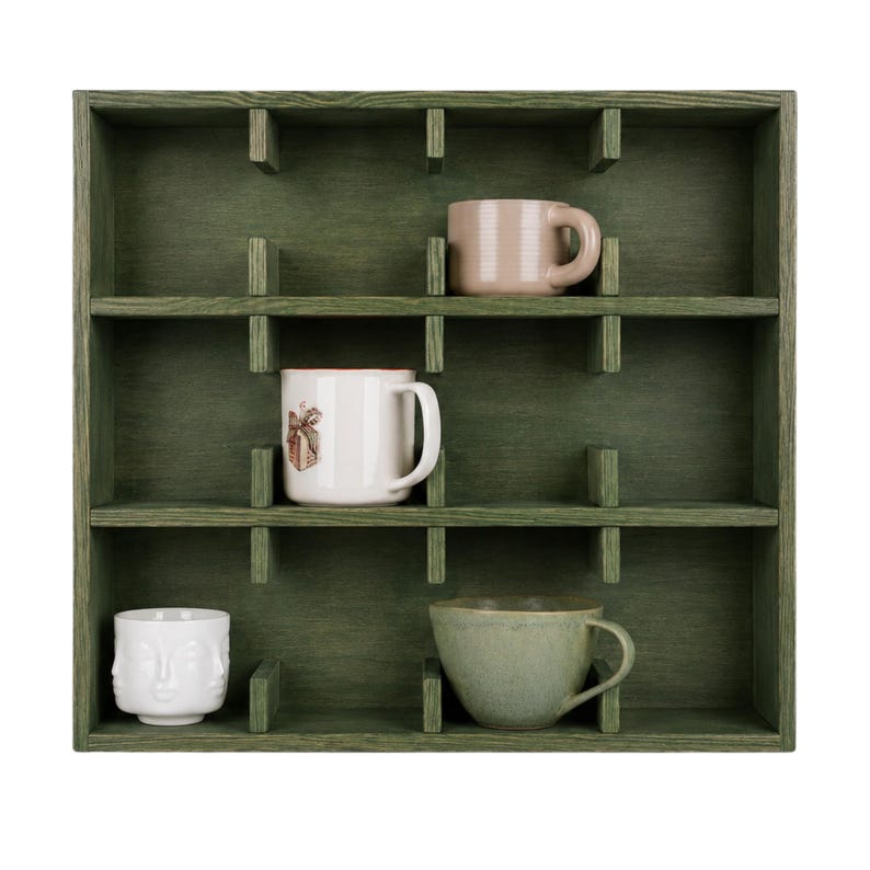 May include: A square, dark green wooden wall shelf with twelve individual compartments. Four mugs are placed inside: a light pink mug, a white mug with a small brown graphic, a white mug with a sculpted face design, and a sage green ceramic bowl.