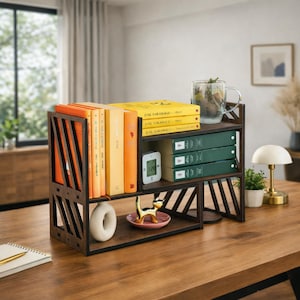 May include: A dark brown wooden desktop bookshelf with an open-frame design featuring diagonal slats. The unit holds several orange and yellow books, three green books, a clear glass mug, a small white digital clock, and a gold cat figurine.