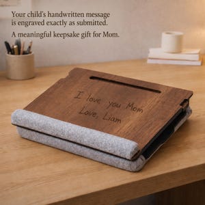 May include: A dark brown wooden tablet or book stand featuring a gray felt base and a horizontal slot. The wood surface is engraved with the handwritten text: I love you Mom, Love, Liam. The stand is angled for viewing and rests on a flat surface.