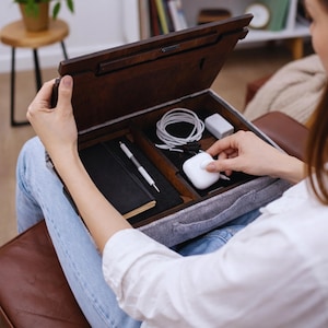 Op de afbeelding: Een houten schootbureau met vakken, open om een zwart notitieboek, pen, oplaadkabels en een witte oplaadcase te onthullen. Het bureau heeft een donkerbruine afwerking en een grijze vilt binnenkant.