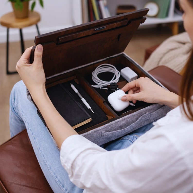 Op de afbeelding: Een houten schootbureau met vakken, open om een zwart notitieboek, pen, oplaadkabels en een witte oplaadcase te onthullen. Het bureau heeft een donkerbruine afwerking en een grijze vilt binnenkant.
