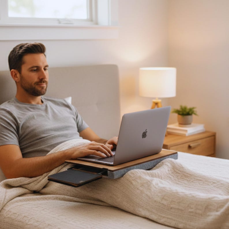 Op de afbeelding: Een persoon gebruikt een zilveren laptop op een houten schoottafel met een grijs kussen. De laptop heeft het Apple-logo. De persoon zit op een bed met een wit dekbed.