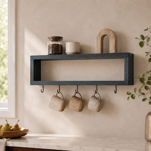 May include: A rectangular dark blue wooden wall shelf featuring five black metal hooks along the bottom edge. Three speckled ceramic mugs hang from the hooks, while a glass jar with dark contents, a small ceramic bowl, and a wooden arch sit on top.