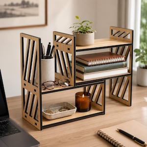 May include: A tiered wooden desk organiser with a light wood finish and black geometric side panels. The unit features two levels holding a ceramic pen holder with pens, spectacles, a small tray with metal clips, a candle, and three stacked notebooks.
