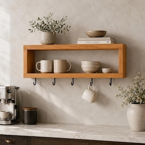 May include: A rectangular light brown wooden wall shelf featuring an open storage compartment and five black metal hooks attached to the bottom edge. The shelf holds two ceramic mugs, two stacked bowls, a small bowl, and a book titled This Is Home.