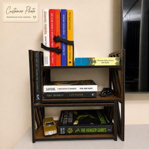 May include: A dark brown wooden bookshelf with three tiers holding various books. The top shelf features a black bookend shaped like two hands holding a row of books. A small gold toy car sits on the bottom shelf, and a small black toy car sits on the middle shelf.