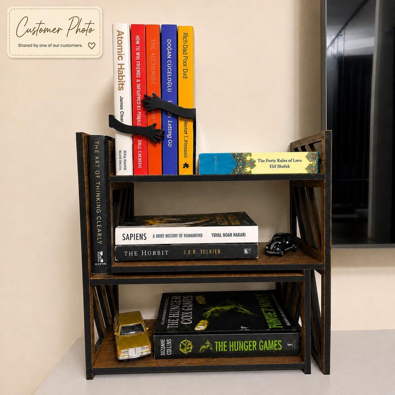May include: A dark brown wooden bookshelf with three tiers holding various books. The top shelf features a black bookend shaped like two hands holding a row of books. A small gold toy car sits on the bottom shelf, and a small black toy car sits on the middle shelf.