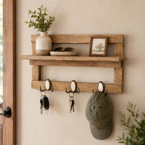 May include: A wooden wall-mounted shelf unit with a shelf, three hooks, and a small shelf. The shelf displays a vase with greenery, sunglasses, and a framed picture. Keys and a hat hang from the hooks. The wood has a natural finish.